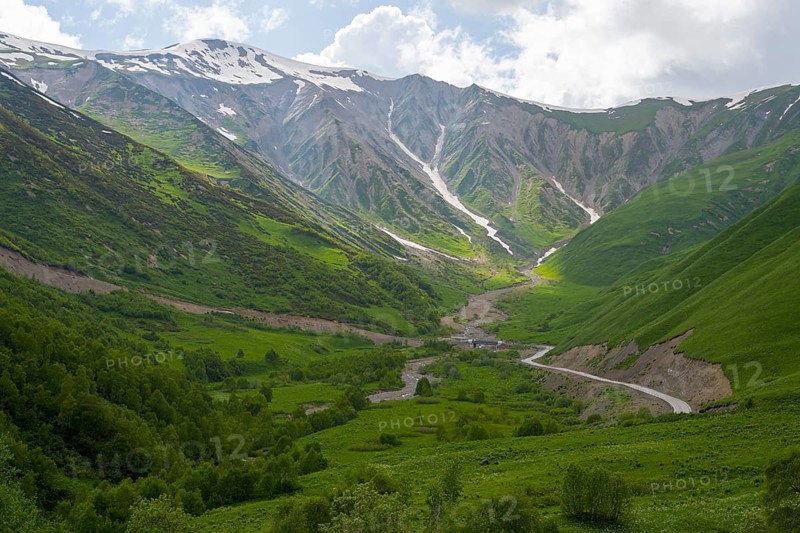 Green valley with snow-covered mountains under cloudy sky and a winding road, near Zagari Pass, Racha-Lechkhumi region and Lower Svaneti, Greater Caucasus, High Caucasus, Georgia
