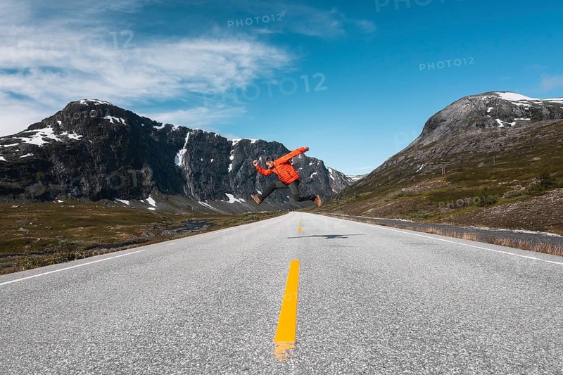 A traveler in a red jacket leaps mid-air on an open mountain road in Norway Framed by dramatic snow-capped peaks and a vibrant blue sky, this scene radiates adventure and freedom