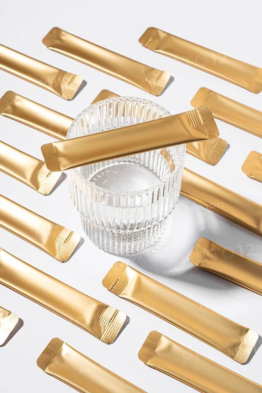 From above an esthetic arrangement of golden jelly collagen sachets with a clear glass resting on a smooth white background, capturing a modern and minimalist style