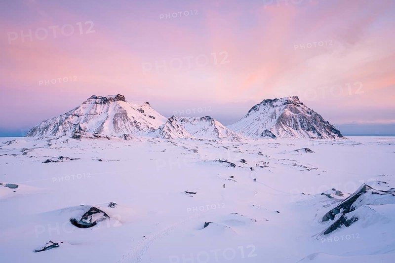 A breathtaking view of Katla Glacier's rugged snow-covered mountains beneath a pastel pink sunrise sky. The untouched snowy plain below adds a sense of serenity and vastness to the arctic scene