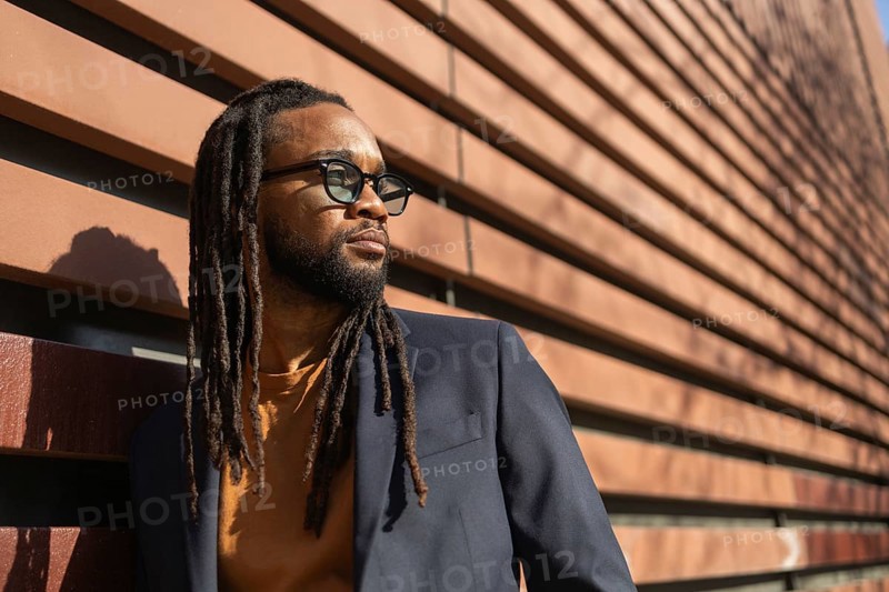 A cuban business man with sunglasses leans against a modern wall in sunlight, showcasing a blend of style and professionalism in an urban setting