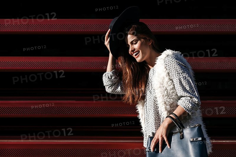 A fashionable woman poses outdoors on a sunny day, wearing a black hat, patterned shirt, and fur vest. She is holding a stylish handbag and smiling joyfully