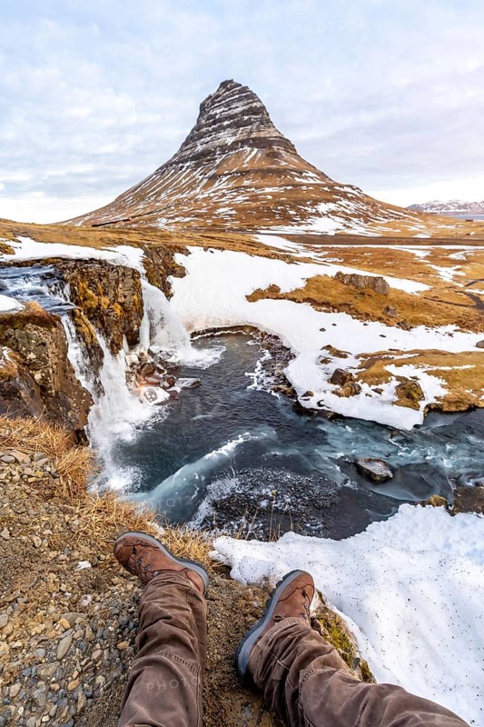 POV of a man sitting on the waterfall with snow on the famous Kirkjufell mountain in winter Iceland