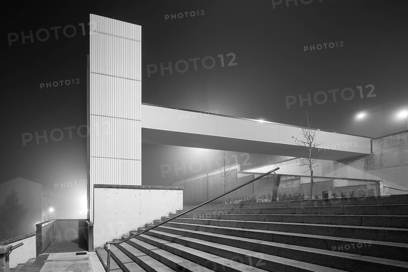 A contemporary urban elevator structure emerges through the dense night fog, creating a surreal atmosphere with its minimalist design and illuminated features