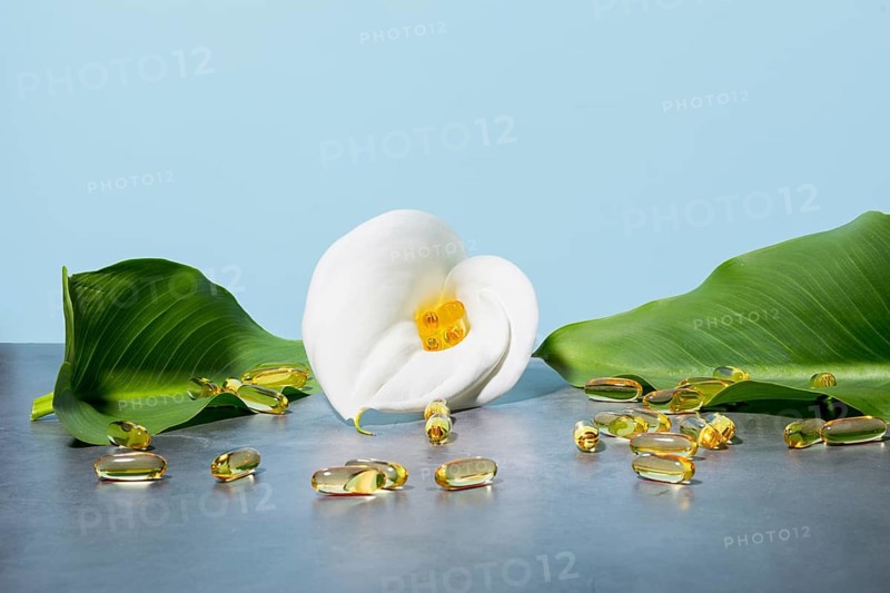 Serene display featuring golden omega-3 oil supplement capsules nestled in a white calla lily, surrounded by lush green leaves against a soft blue background, emphasizing beauty and wellness