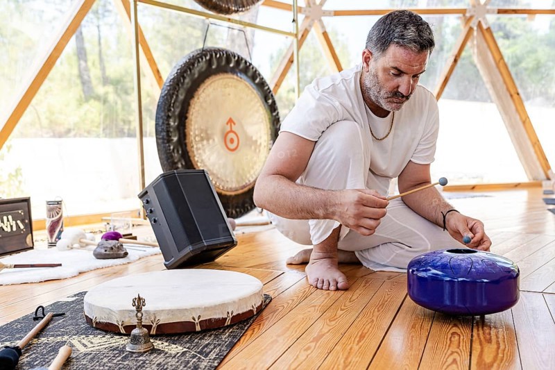 A male sound therapist conducts a sound therapy session with steel tongue drum, and other instruments including the a large gong in a serene wooden room, promoting relaxation and wellness