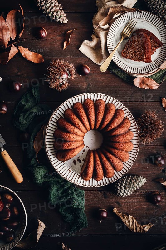 A delicious chestnut cake sits elegantly on a rustic table, surrounded by autumn leaves and pine cones. Its rich brown color reflects the cozy seasonal atmosphere perfectly