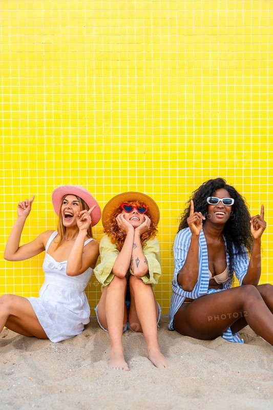Three diverse women wearing summer clothes and hats are sitting on the sand, pointing upwards with their index fingers, in front of a yellow tiled wall