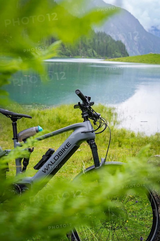 An e-bike is parked on the lakeshore. The surrounding nature is lush and green, and the mountains can be seen in the background: Stilluptal, Zillertal. Austria