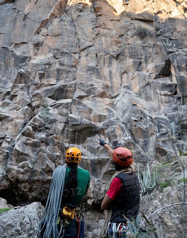 Two climbers equipped with helmets and ropes assess a towering rock face for a traditional climbing route in a natural setting, showcasing their adventurous spirit