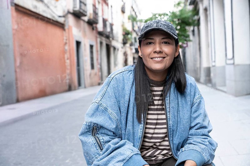 Latina woman wearing a striped shirt and denim jacket smiles confidently while sitting on a vibrant urban street lined with colorful buildings and lush greenery