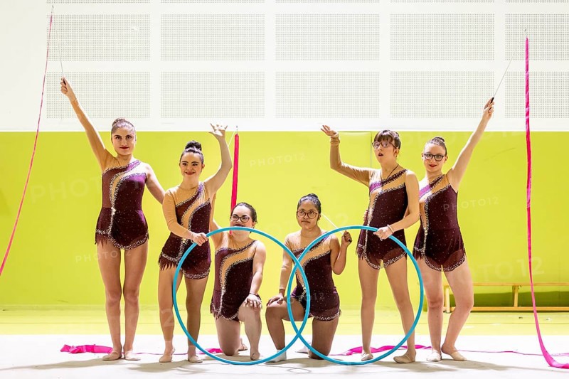 Group of gymnasts whit disability in maroon outfits performing with hoops and ribbons in a brightly lit arena. They showcase elegance, and athleticism, creating a captivating display and inclusive