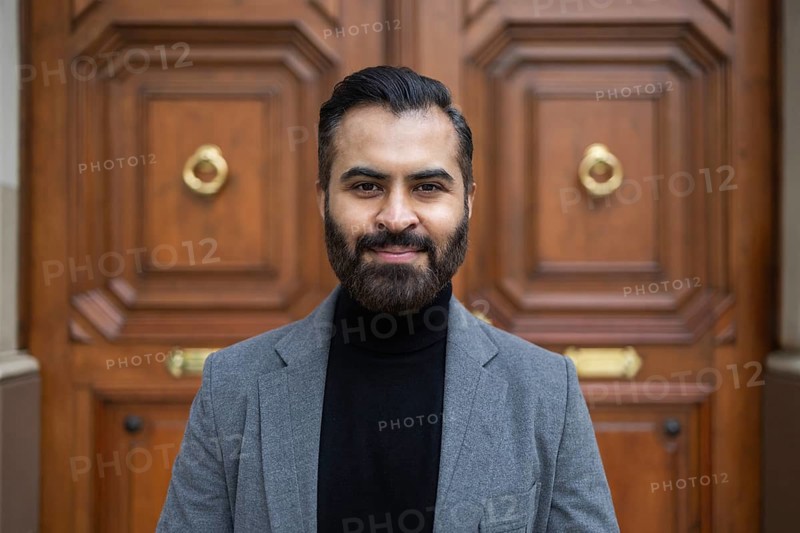 Portrait of a confident Indian businessman in a grey coat and black turtleneck, standing in front of a classic wooden door, looking directly at the camera