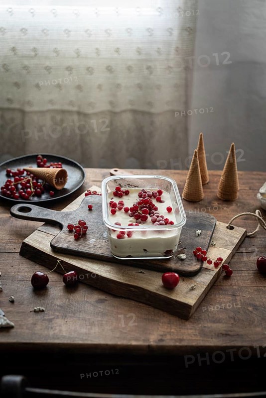 Rustic scene of homemade vanilla ice cream garnished with red currants and berries, served on a wooden board with scattered cherries