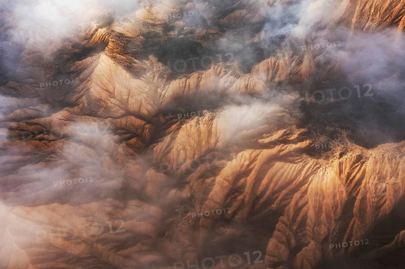 Captivating aerial view showcasing majestic mountain peaks partially enveloped in soft, swirling clouds The rugged terrain is highlighted by warm sunlight, creating a mesmerizing contrast