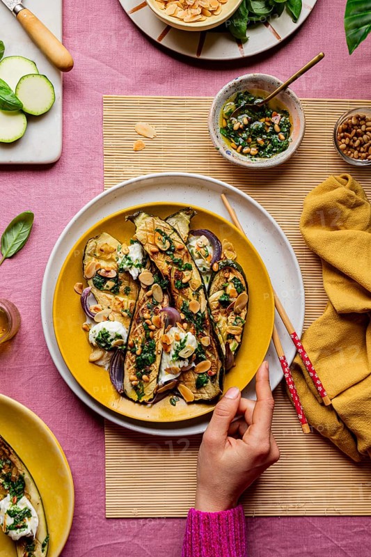 Top view of cropped unrecognizable hand serving a beautifully garnished roasted courgettes served on a vibrant yellow plate, alongside fresh ingredients and a savory dressing, showcasing a colorful and tempting presentation