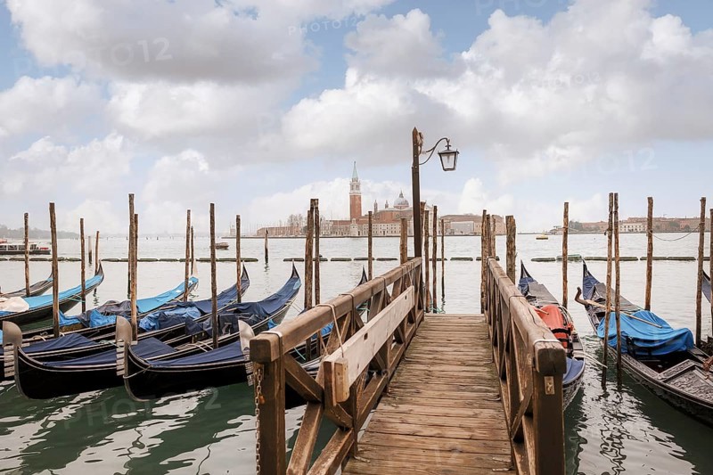 Scenic view of a wooden dock leading to gondolas in Venice, Italy. The background showcases the iconic skyline and gentle clouds enhancing the serene atmosphere