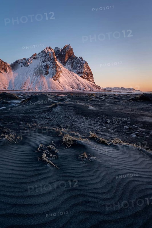 A stunning view of Stokksnes Mountain in Iceland, illuminated by the warm light of sunrise. The dark sand and icy terrain create a dramatic and serene winter scene
