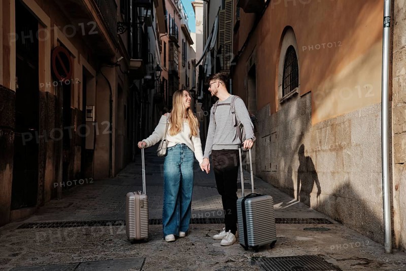 A young couple walks hand in hand down a narrow street, luggage in tow, as they search for their dream vacation home in the picturesque town of Calatrava, Majorca, Spain