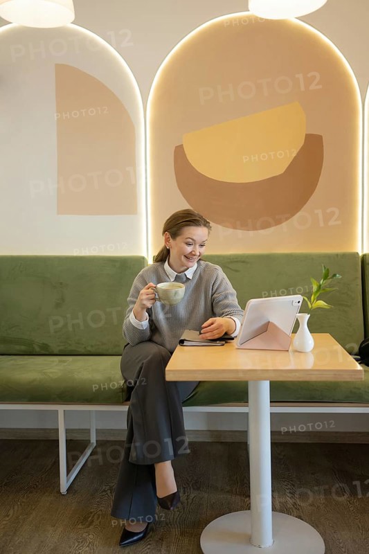 A woman relaxes in a stylish cafe, sipping coffee and using a tablet. The tranquil setting is enhanced by modern decor and soft lighting, creating a cozy ambiance