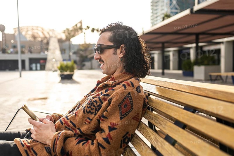 A man sits calmly on a bench outdoors with a smartphone Wearing sunglasses and a stylish patterned sweater, he enjoys the peaceful urban environment in the sunlight
