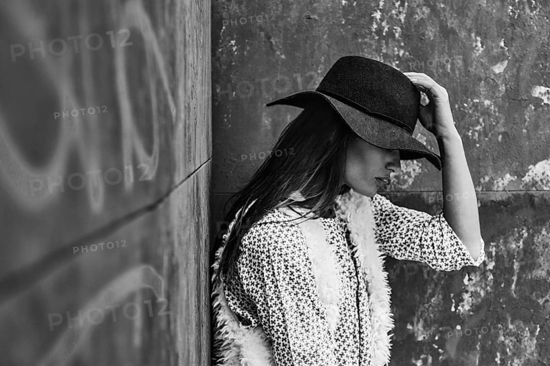 A black and white image of a woman in a hat resting against a textured wall. She wears a stylish outfit with a patterned top and fur vest, exuding a sense of calm and reflection