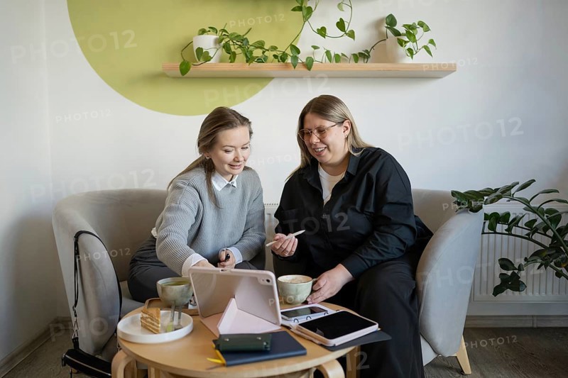 Two women sit on cozy chairs in a cafe, sharing conversation over coffee. A tablet rests on the table surrounded by notebooks, enhancing the relaxed setting