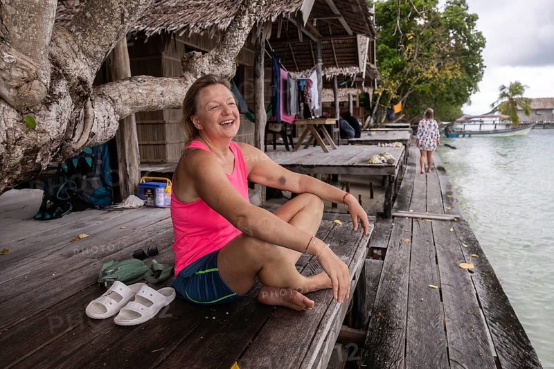 A joyful mature Caucasian woman tourist relaxes on a wooden dock at a rustic homestay, surrounded by the tranquil waters of Kri Island in the Raja Ampat Archipelago, West Papua, Indonesia