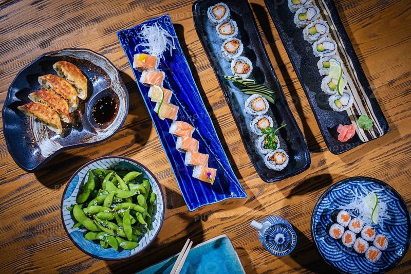 Overhead view of a colorful assortment of japanese cuisine, including sushi rolls, gyoza, edamame, and dipping sauce, served on a rustic wooden table