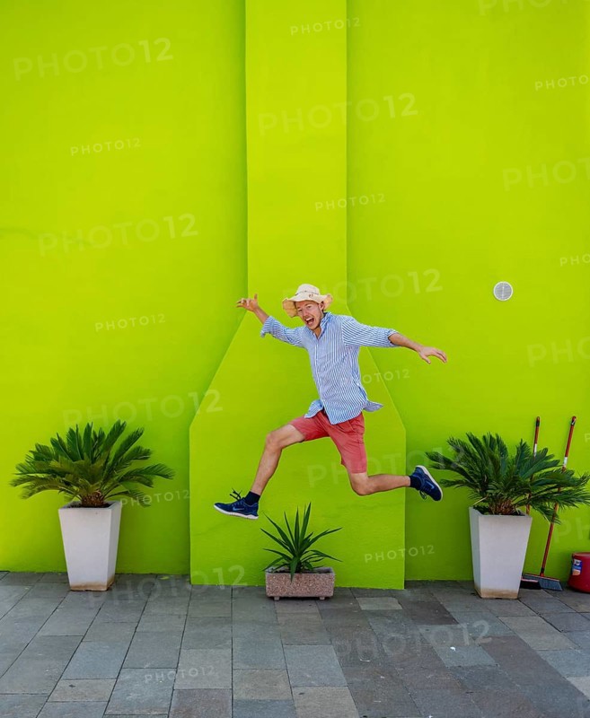 Young man jumping over plant pot in front of green house wall, green house facade with flowers, colourful houses on the island of Burano, Venice, Veneto, Italy