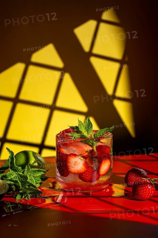 A refreshing strawberry mojito garnished with fresh mint and sliced strawberries in a clear glass. Set on a vibrant red surface, with a lime and mint leaves beside it against yellow background