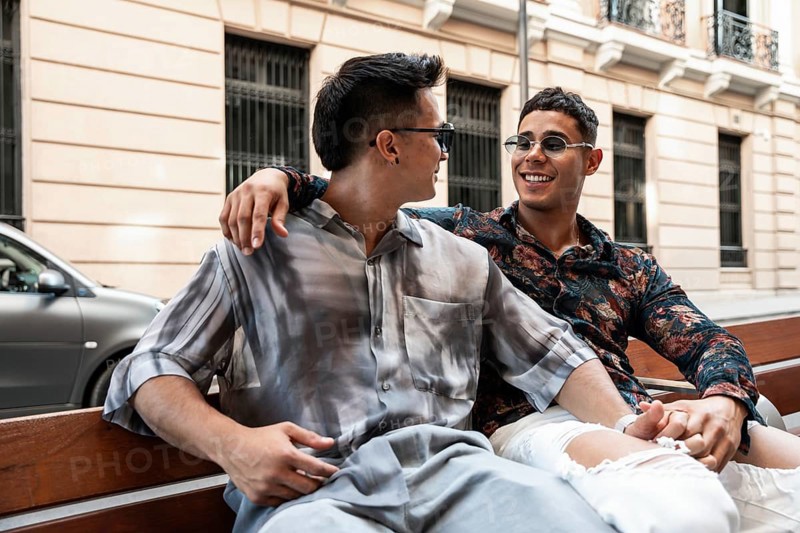 A cheerful latin gay couple sits close together on a bench, laughing and enjoying a sunny day in the vibrant streets of Madrid, showcasing love and happiness