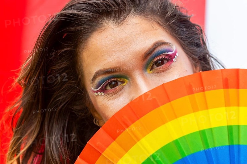 A woman with vibrant rainbow makeup partially conceals her face with a pride-themed fan. Her expressive eyes convey the spirit of inclusion, celebrating LGBTQIA+ identity
