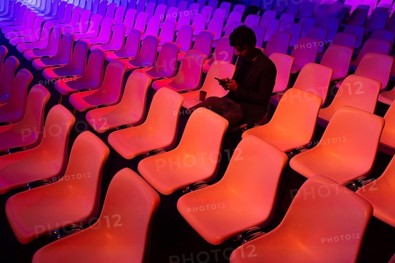 A indian man sits among vibrant, colorful auditorium chairs at a conference of AI in Amsterdam engrossed in his smartphone. The scene contrasts solitary focus against a backdrop of empty seats