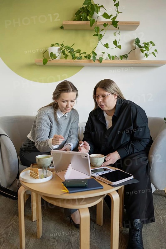 Two women engage in a collaborative discussion at a cozy cafe, using a tablet. They sit at a wooden table with coffee cups, surrounded by greenery and a relaxed atmosphere