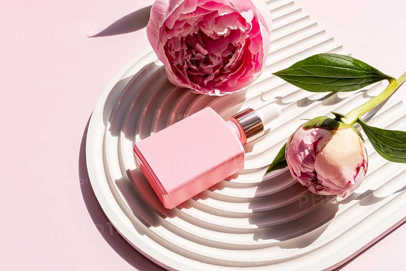 A pink cosmetic bottle is elegantly placed on a white decorative tray, surrounded by beautiful pink peonies. The sunlight enhances the subtle aesthetics of the scene