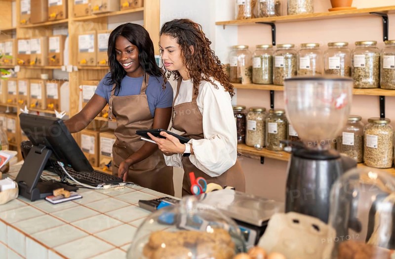 Two clerks at an organic supermarket at the counter. They smile and engage with the cash register and a digital tablet, surrounded by jars of natural products