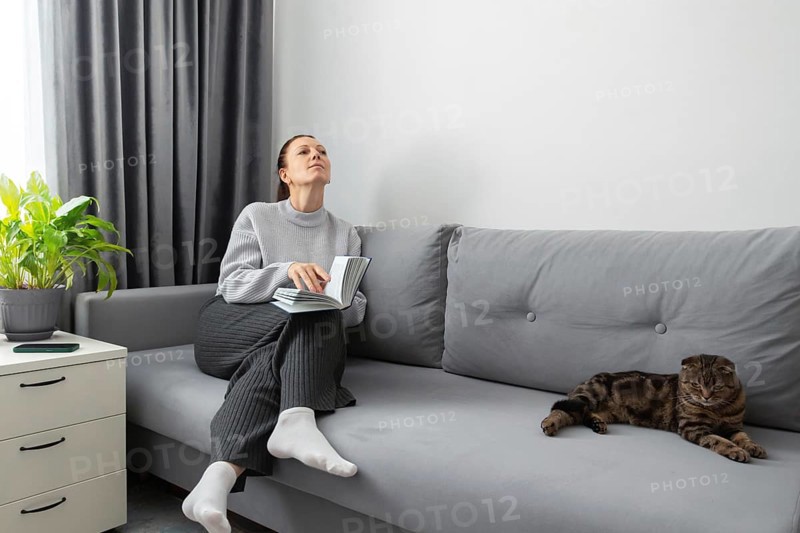 A woman sits on a gray sofa, holding a book, looking peaceful and contemplative. Beside her, a cat lounges calmly. A green plant and drawer add to the room's tranquility