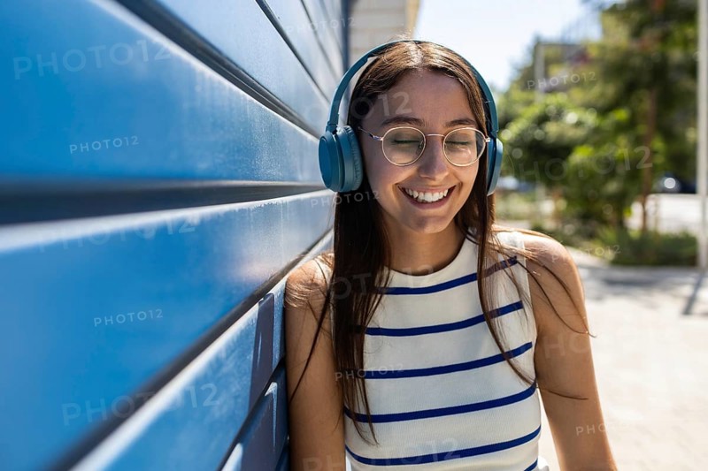 Young woman enjoying her urban lifestyle outdoors, leaning against a blue wall while listening to music through headphones. Her eyes are closed, and she is smiling peacefully