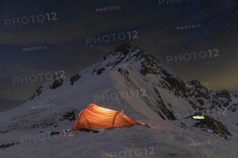 A glowing tent in a snowy mountain landscape under a starlit sky. Perfect for ski touring and winter adventures, this serene setting captures nighttime wilderness beauty