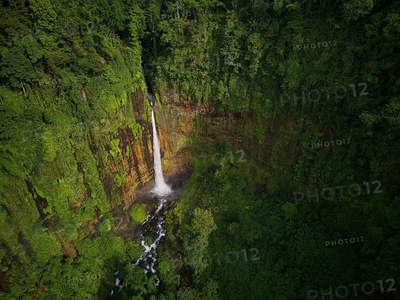 Aerial shot capturing the majestic Kaspa Biru Waterfall descending into a natural gorge, surrounded by dense tropical rainforest. The waterfall creates a serene atmosphere as it flows through the verdant landscape