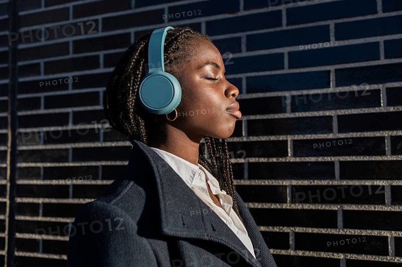 An African American businesswoman with braids enjoys a break, listening to music through headphones, eyes closed in relaxation, leaning against a brick wall outdoors