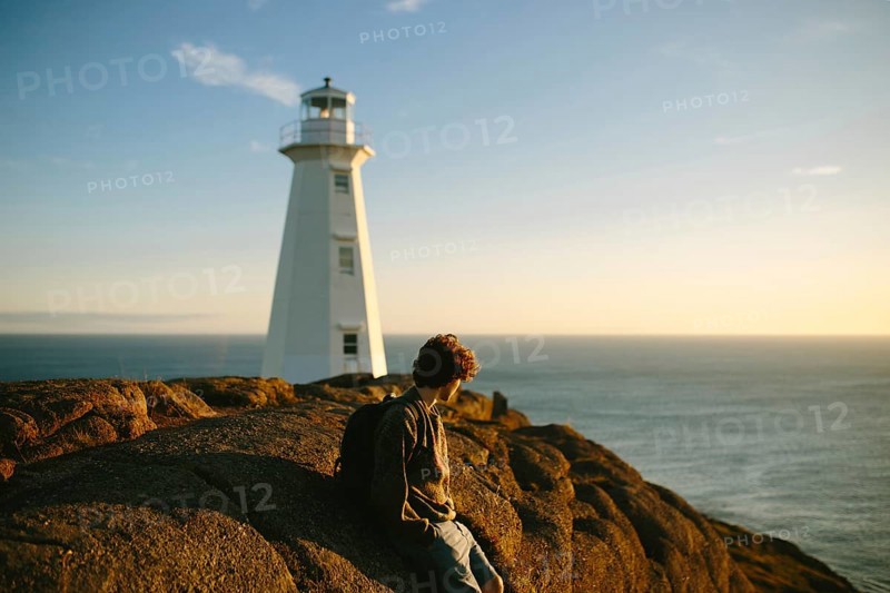 Side view of unrecognizable young man sitting on rocks gazing at the ocean near Cape Spear Lighthouse at sunset. The serene environment highlights a tranquil moment at this historic site in Newfoundland & Labrador, Canada