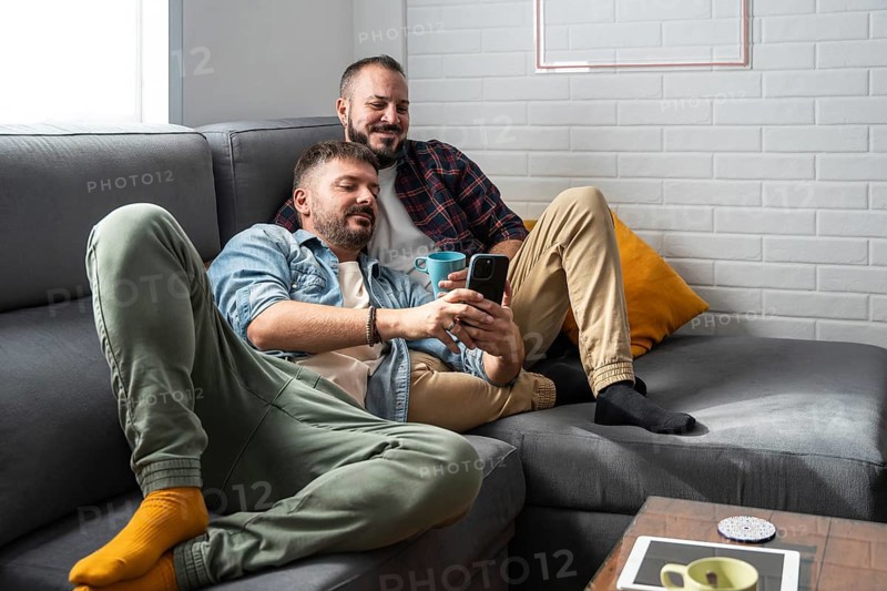 A content gay couple lounges on a comfortable gray sofa, enjoying coffee together while browsing a smartphone. They rest casually against soft pillows, sharing a peaceful moment