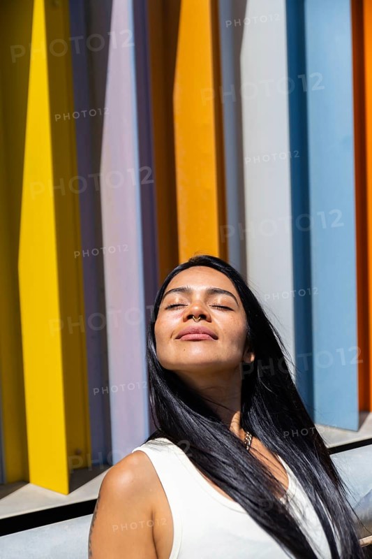 A woman in a colorful outfit enjoys the sunlight, standing in front of a backdrop of vibrant, modern architecture. Her serene expression contrasts with the urban vibrancy