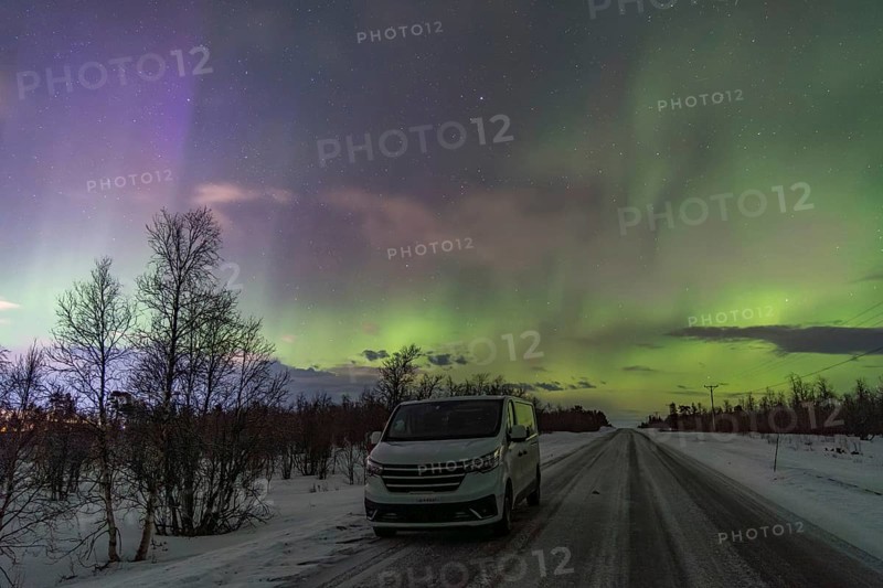 A mesmerizing display of Aurora Borealis fills the sky above a snowy road with a vehicle parked facing the viewer near Kiruna, Sweden. The winter scene captures the vibrant greens and purples of the northern lights