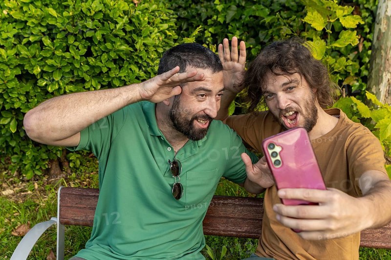 Two friends have fun taking a playful selfie while sitting on a park bench The man in green, who is blind, smiles and poses enthusiastically alongside his friend, celebrating their joyful bond