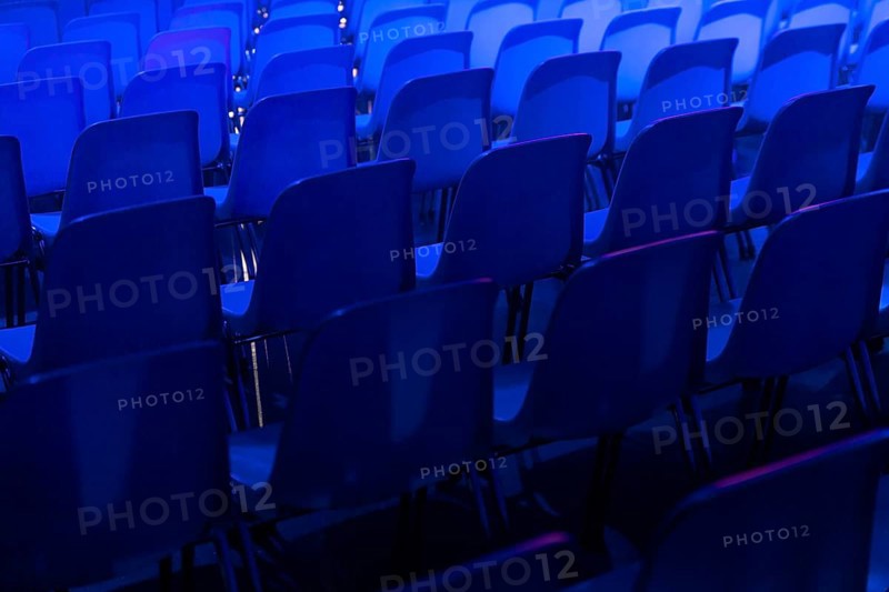 A series of empty blue chairs are arranged in neat rows under a dramatic blue spotlight at a conference of AI in Amsterdam, creating a cool and contemporary atmosphere