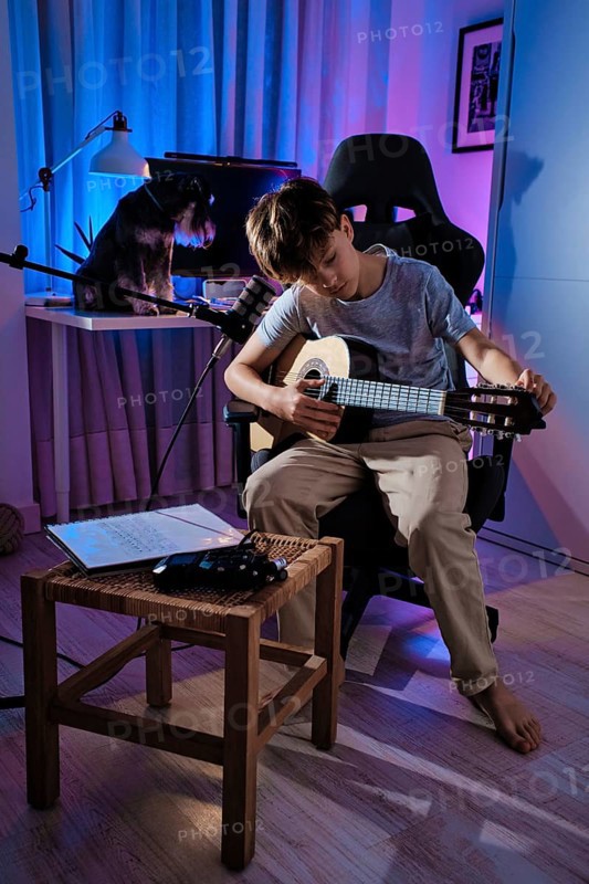 A young boy tuning his guitar in a cozy, blue lit room, with a microphone and sheet music nearby. A dog rests on a desk behind him, creating a serene atmosphere