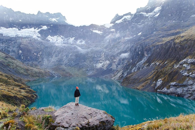 A woman stands on a rocky ledge, overlooking the stunning turquoise lake and surrounding peaks of El Altar volcano in Ecuador, embodying the spirit of adventure